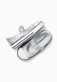 Zapatos de lona plateados con lentejuelas, con una correa de hebilla, punta de goma blanca y suela a rayas en blanco y negro. Silueta achatada.