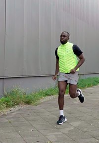 Gilet imbottito verde neon, maniche corte nere, pantaloni grigi, scarpe da corsa nere, che corre lungo un marciapiede accanto a un'area verde.