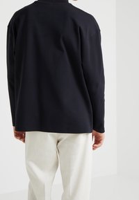 Schwarzes langärmliges Hemd mit lockerem Schnitt, hohem Kragen und glatter Textur, kombiniert mit hellfarbenen Hosen.