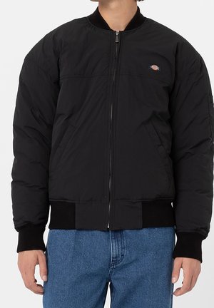 Person trägt eine schwarze Bomberjacke mit Reißverschluss, gerippten Bündchen und Saum, kombiniert mit blauen Jeans aus Denim.