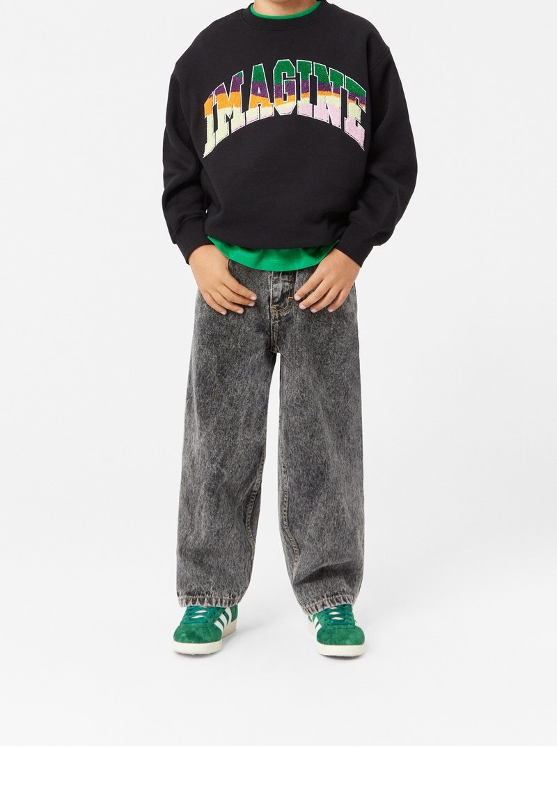 Enfant portant un sweat-shirt noir avec le texte coloré "IMAGINE", une chemise verte en dessous, un jean gris ample et des baskets vertes avec des bandes blanches.