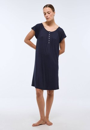Femme debout pieds nus dans une robe bleu marine à manches courtes avec de petits pois blancs et des boutons, regardant vers le bas avec les mains dans le dos.