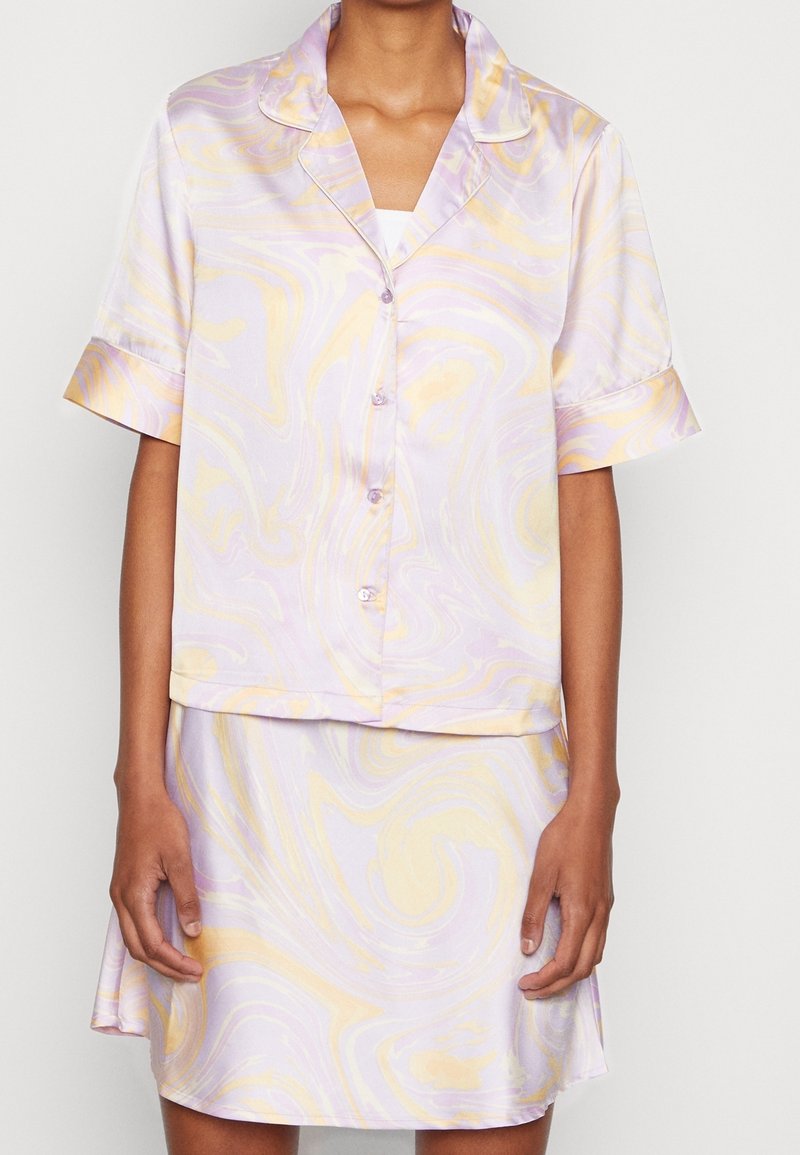 Femme portant une chemise boutonnée à manches courtes marbrée lavande pastel et jaune avec une jupe assortie sur un fond uni.