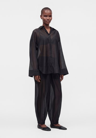 Personne portant une chemise noire ample et transparente à boutons et un pantalon transparent assorti avec des poignets élastiques, debout et faisant face à l'avant sur un fond uni.
