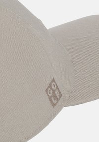 Casquette beige en tissu texturé, avec un patch logo brodé marron. La visière courbée est bien dessinée et soigneusement définie.