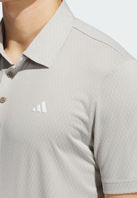 Vaaleanharmaa polo-paita, jossa on teksturoitu kangas, kolme nappia ja valkoinen logo. Lyhyet hihat ja klassinen kaulustyyli.