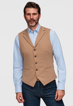 Mand med skæg iført lyseblå skjorte, beige knappet vest og mørkeblå jeans, stående mod en ensfarvet lys baggrund.