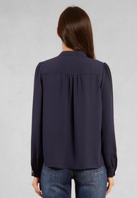 Blusa a maniche lunghe blu navy con colletto a bottoni, dettaglio plissettato sul retro e vestibilità sagomata. Tessuto morbido, leggero.