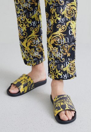 Jambes portant un pantalon noir avec des motifs baroques dorés et le texte "Versace Jeans Couture", associées à des sandales slides noires et dorées assorties.
