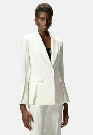 Donna che indossa un blazer bianco su misura e pantaloni abbinati con una maglietta nera sotto, in piedi davanti a uno sfondo semplice.