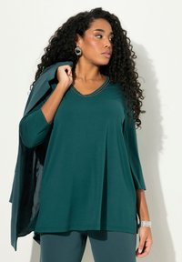 Tumman turkoosi V-pääntie pusero, jossa kolmen neljänneksen hihet, pehmeää kangasta ja hopea-omaan reunus kaula-aukolla. Yhdistetty yhteensopiviin housuihin ja takkiin.