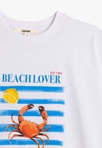 Biała bawełniana koszulka z centralnym motywem w postaci pomarańczowego kraba, niebieskich pasków, żółtej cytryny oraz napisem "BEACH LOVER" w niebieskim i czerwonym kolorze.