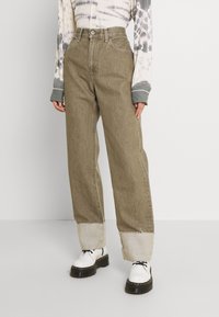 Jeans anchos de color verde oliva claro con dobladillo deshilachado, diseño de cintura alta, trabillas para cinturón y detalles de costura, combinados con zapatos de plataforma blancos.