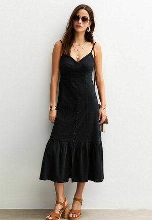 Vestido midi negro de mezclilla, con escote en V, botones en la parte delantera, dobladillo acampanado y tirantes finos. Combinado con sandalias de tacón marrones.