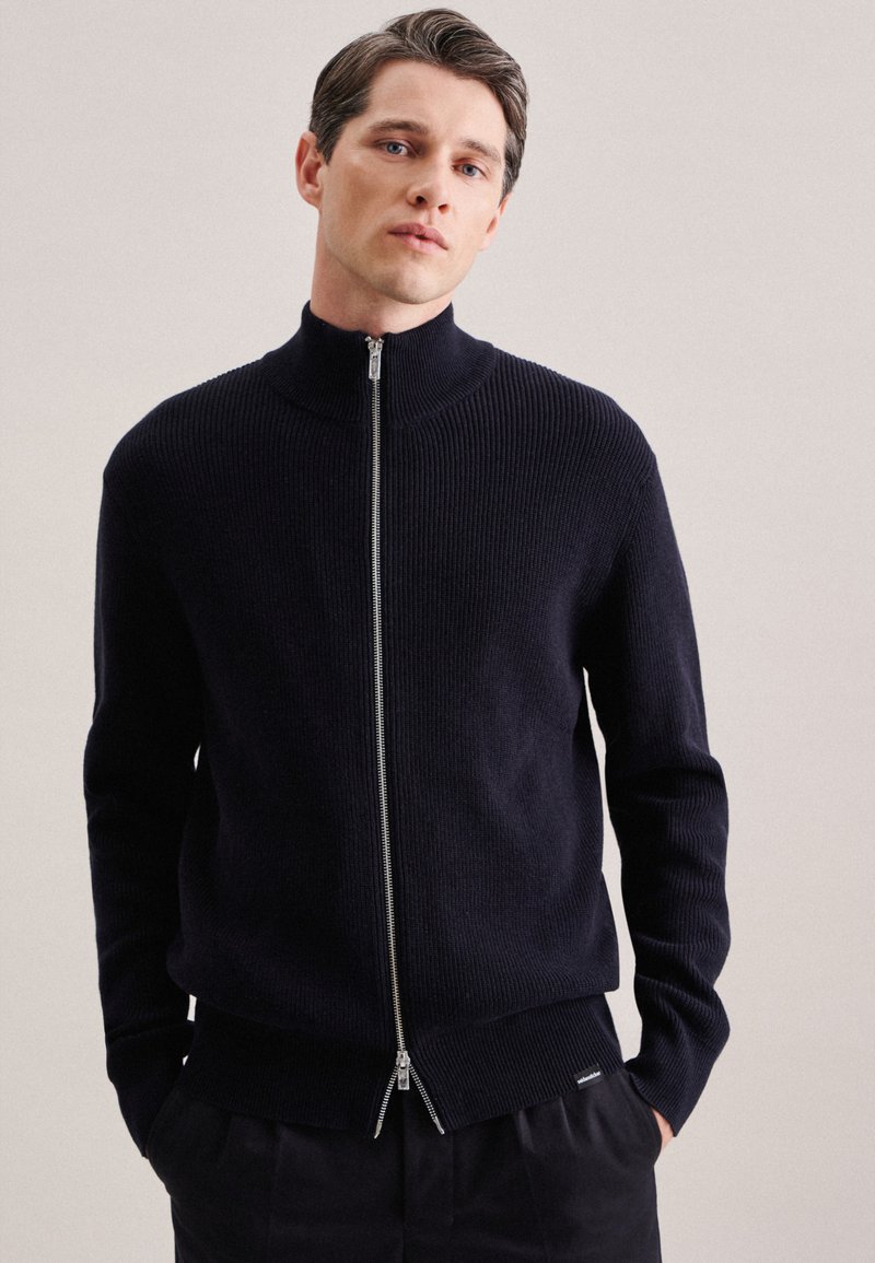 Sweat à fermeture éclair navy avec un col montant, texture côtelée et fermeture éclair argentée. Présente des poignets et un ourlet ajustés, associé à un pantalon sombre.