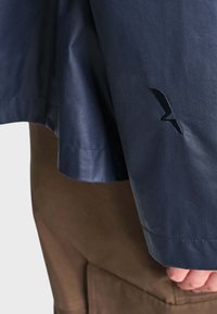 Manche de veste bleu foncé avec un petit logo noir au-dessus du poignet, portée sur un pantalon marron avec des coutures de poche visibles.