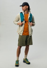 Veste beige légère, chemise orange, shorts verts et chaussures de randonnée colorées. Comprend un sac à dos bleu et une casquette noire.