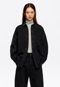 Zwarte denimjas met gestifte accenten langs de schouders, knopen aan de voorkant, gecombineerd met een grijze turtleneck en zwarte wijde broek.