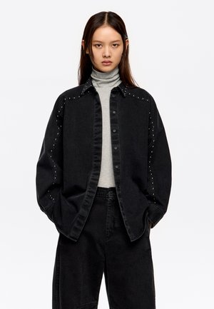 Czarna kurtka dżinsowa z ćwiekowanymi akcentami na ramionach, zapinana na guziki, w zestawieniu z szarym golfem i czarnymi spodniami typu wide-leg.