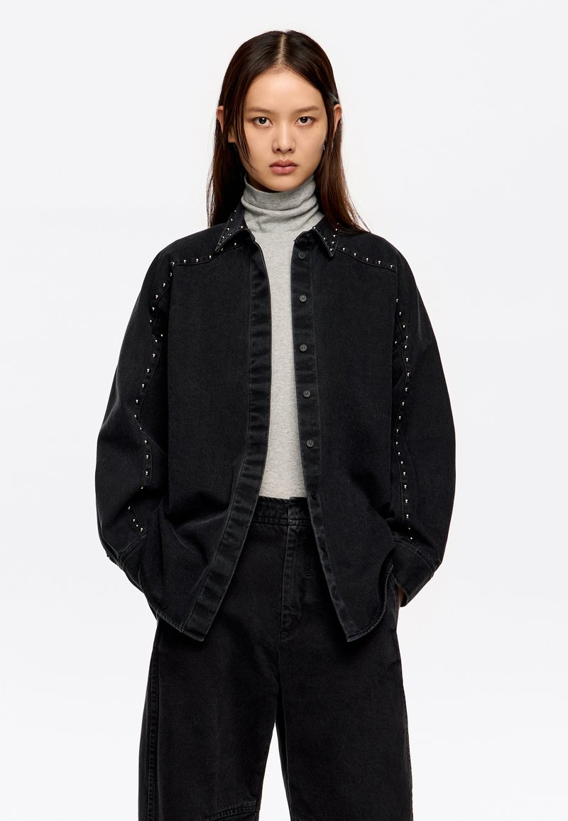 Zwarte denimjas met gestifte accenten langs de schouders, knopen aan de voorkant, gecombineerd met een grijze turtleneck en zwarte wijde broek.