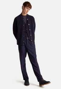 Cardigán de canalé azul marino con botones de presión sobre una camisa azul marino oscura, combinado con pantalones de vestir azul marino y zapatos de ante negros con acentos naranjas.