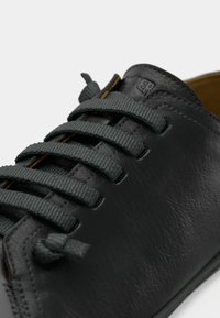 Musta nahkakenkä harmailla, kuvioiduilla nauhoilla, sileällä pinnalla ja pelkistetyllä muotoilulla. Kenkässä näkyvä logo kieliosassa ja kontrastitikkaus.