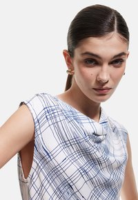Weißes Oberteil mit blauem Karomuster, Schalkragen und ärmellosem Design. Glatte Stoffstruktur mit seitlichen Ausschnitten für einen modernen Look.