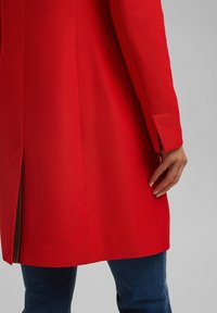 Rode textuurrijke jas met een getailleerde pasvorm, een gesplitste achterkant en scherp gesneden mouwen. Draagt over een blauwe spijkerbroek, met een modern ontwerp.