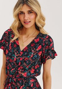 Žena s vlnitými blond vlasy, oblečená v černých květinových šatech s červenými květy, zlatými kruhovými náušnicemi a kulatým zlatým náhrdelníkem.