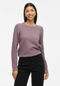 Pull en tricot couleur mauve avec un motif de maille texturé, col rond et manches longues, associé à un pantalon noir.