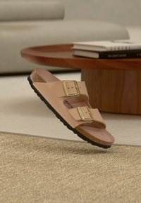 Μπεζ σανδάλι με δύο λουριά, χρυσά αγκράφες και φελλό σόλα που αιωρείται πάνω από μπεζ υφασμάτινο χαλί, δίπλα σε ξύλινο τραπεζάκι με βιβλία.