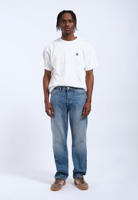 T-shirt bianca in cotone con una piccola grafica, abbinata a jeans di denim blu chiaro e sneaker grigio-marroni. Design minimalista, vestibilità rilassata.