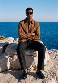 Uomo che indossa una giacca trapuntata marrone, occhiali da sole neri, pantaloni neri e scarpe, seduto su scogliere rocciose vicino all'oceano sotto un cielo limpido.
