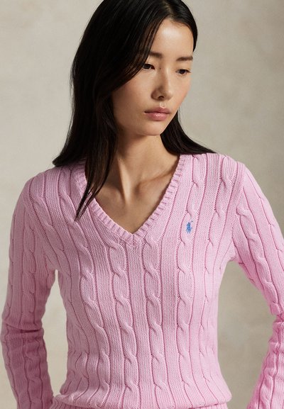 Frau mit langem dunklem Haar, die einen pinkfarbenen, kabelgestrickten Pullover mit V-Ausschnitt und kleinem gesticktem Logo auf der Brust trägt und nach unten schaut.