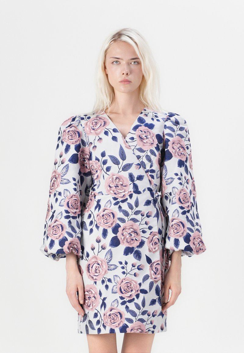 Φλοράλ φόρεμα τύπου wrap με λευκή βάση, μεγάλες ροζ τριανταφυλλιές και ναυτικές φύλλα. Τόλμηρες, φουσκωτές μανίκες και πλεξούδα στη μέση. Λείο ύφασμα.