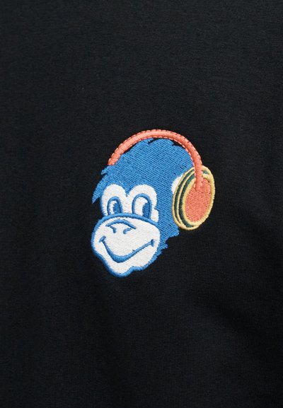 T-shirt en tissu noir avec un singe bleu souriant brodé, portant un casque orange. La couture détaillée met en valeur le design.