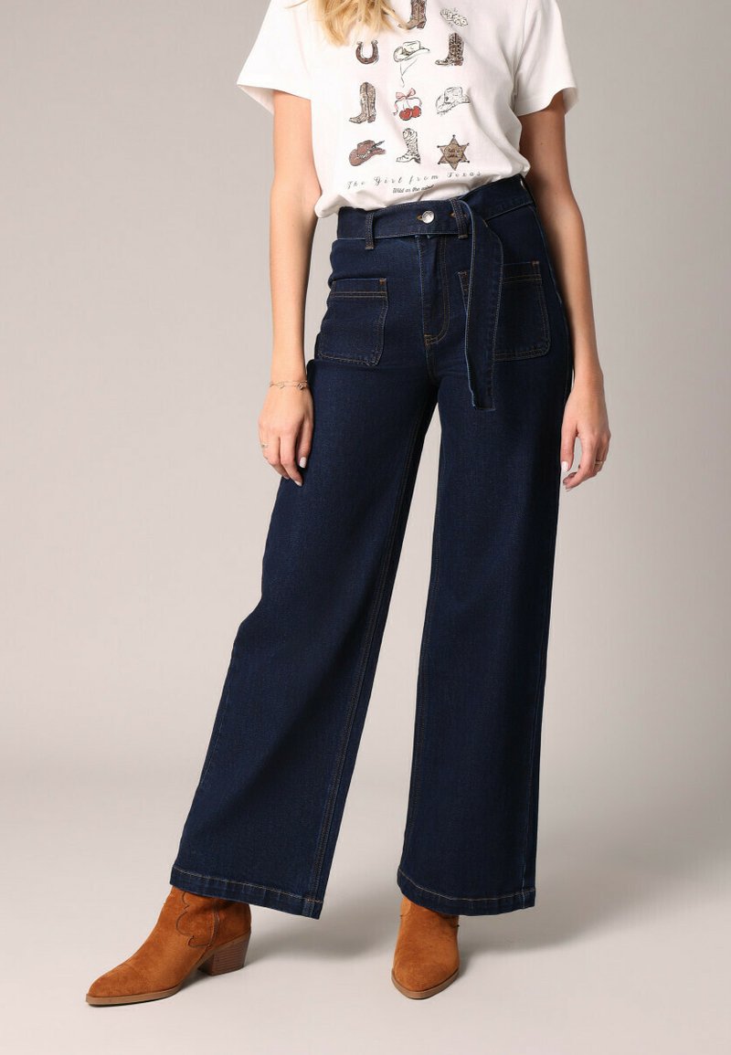 Wijd-uitlopende donkere denim jeans met een hoge taille, riemlussen en voorzakken. Gecombineerd met een wit graphic t-shirt en bruine suede enkellaarzen.