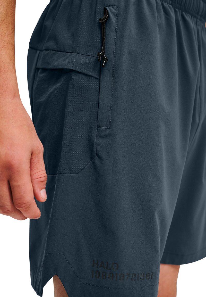 Nærbillede af en hånd ved siden af marineblå shorts med elastisk taljebånd, lynlåslynlomme og svag sort tekst "HALO 196819721981" nær kanten.