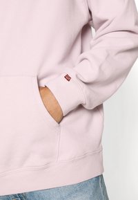 Sudadera rosa clara hecha de una suave mezcla de algodón. Presenta un bolsillo tipo canguro y una pequeña etiqueta de logo roja en el puño.