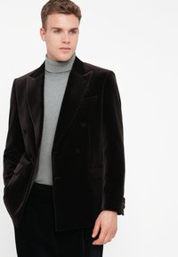 Jovem vestido com um blazer preto de veludo duplo sobre um suéter de gola alta cinza claro, posando contra um fundo branco.