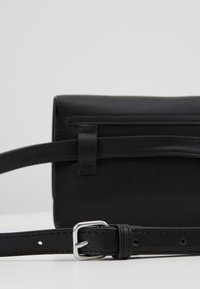 Sac bandoulière en cuir noir de forme rectangulaire, avec rabat à pli, sangle réglable et bouclerie en métal argenté.