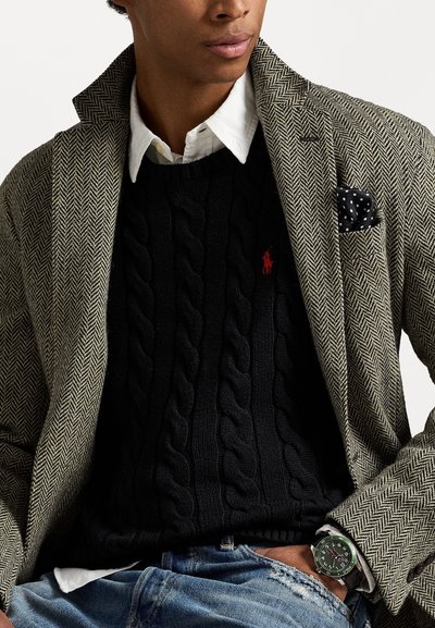 Blazer en motif chevrons beige, pull noir en mailles torsadées, chemise blanche à col, pochette à pois noirs, montre verte et noire.