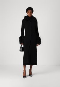 Zwarte ribbelige lange jas met een gevederde kraag en manchetten, knopen aan de voorkant en een rechte silhouet, gecombineerd met zwarte hakken en een kleine tas.