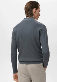 Grau gerippter Strickpullover mit langen Ärmeln und Stehkragen, kombiniert mit hellbeigen Hosen, von hinten gezeigt.