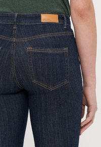 Jeans in denim blu scuro con una texture liscia, caratterizzati da una patta in pelle in vita e due tasche posteriori con cuciture a contrasto.