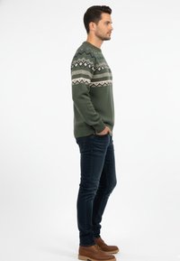 Jersey verde de punto con patrones geométricos en beige y verde oscuro, combinado con vaqueros oscuros y botas marrón claro, de pie de lado.