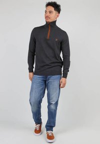 Sweat à fermeture éclair gris foncé avec accents orange, col côtelé et logo ; associé à un jean bleu clair et des baskets brunes. Texture lisse.