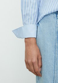 Chemise bleu clair rayée avec un large poignet, associée à un denim clair. Le tissu semble lisse avec un léger éclat.