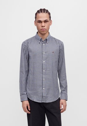Un homme aux cheveux tressés se tient face à l’objectif, vêtu d’une chemise boutonnée à carreaux bleu marine et blancs et d’un pantalon foncé, sur un fond uni.