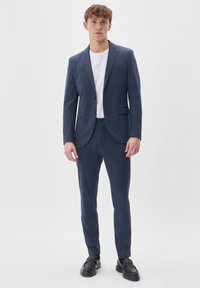 Navy blue suit made of textured fabric, featuring a single-button blazer with lapels and slim-fit trousers, paired with a white t-shirt and black shoes.